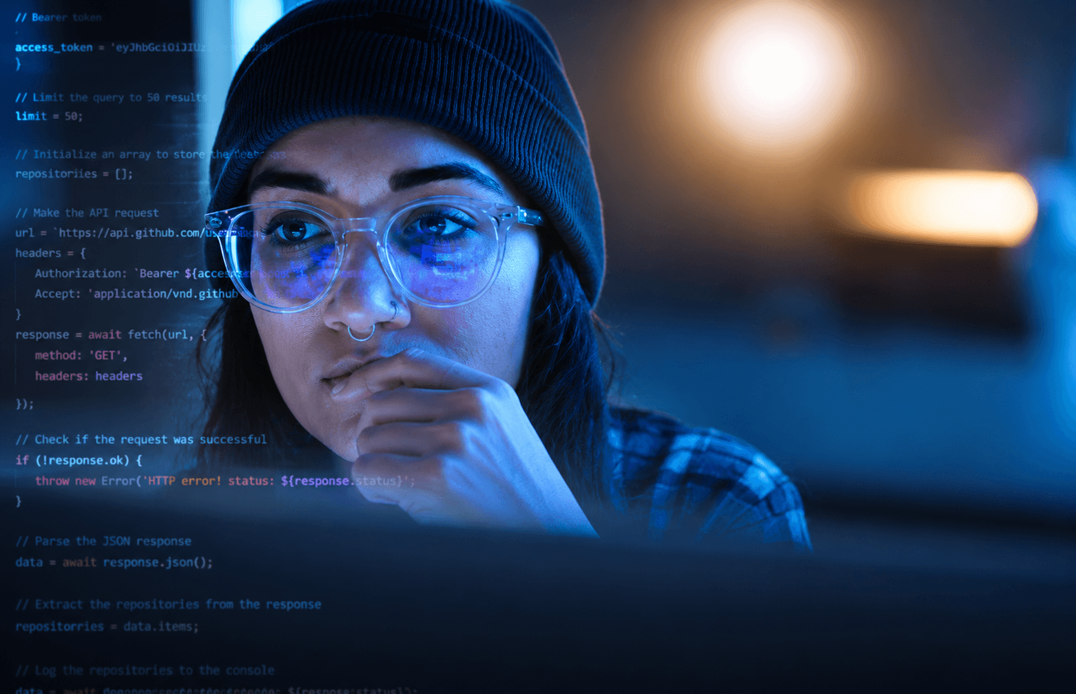 Woman in glasses and beanie intently coding on a computer screen with blue light.