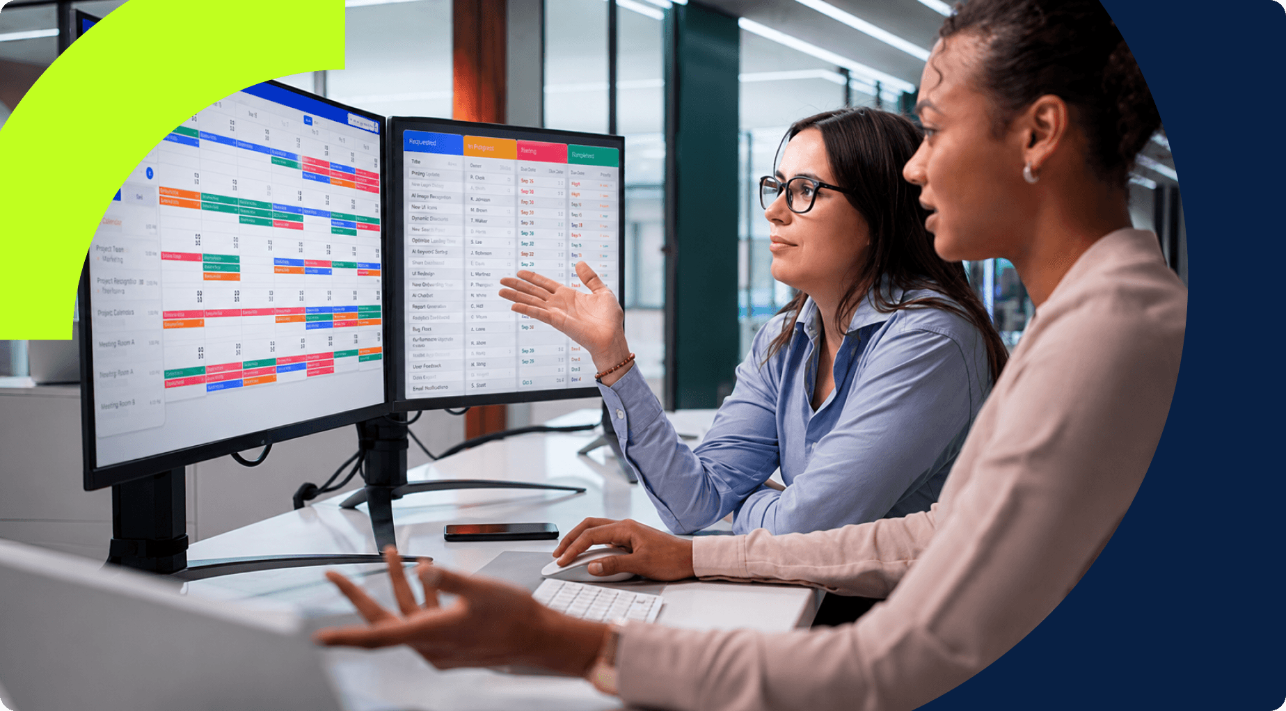 Two women collaborate, discussing project schedules displayed on dual computer monitors.