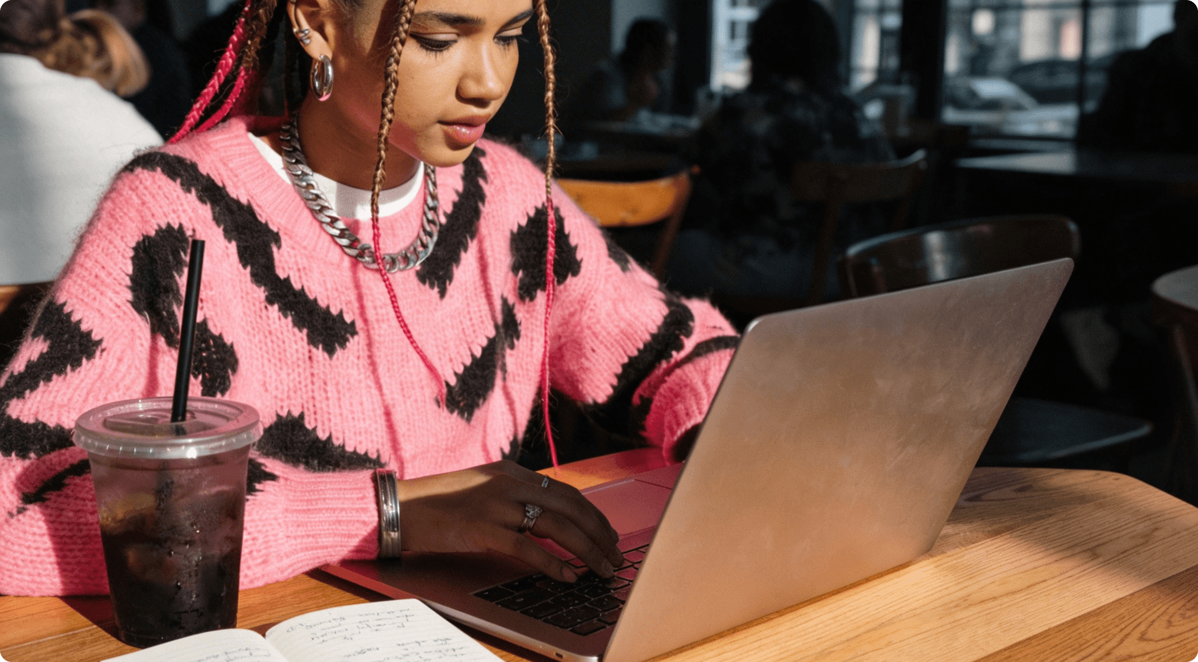 Young woman in a pink and black sweater types on a laptop in a busy cafe.