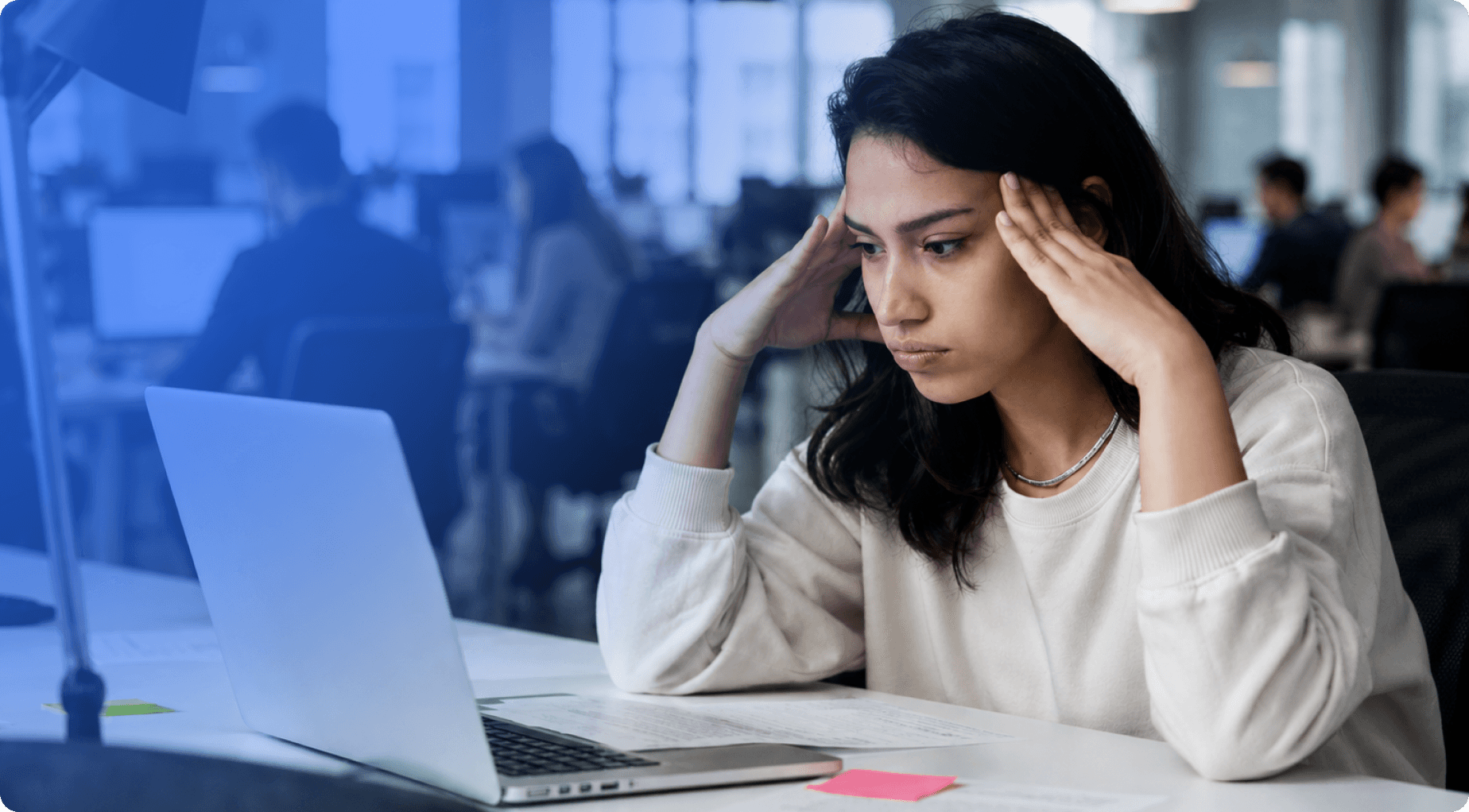 A stressed young woman holds her head while looking at a laptop in a busy office.