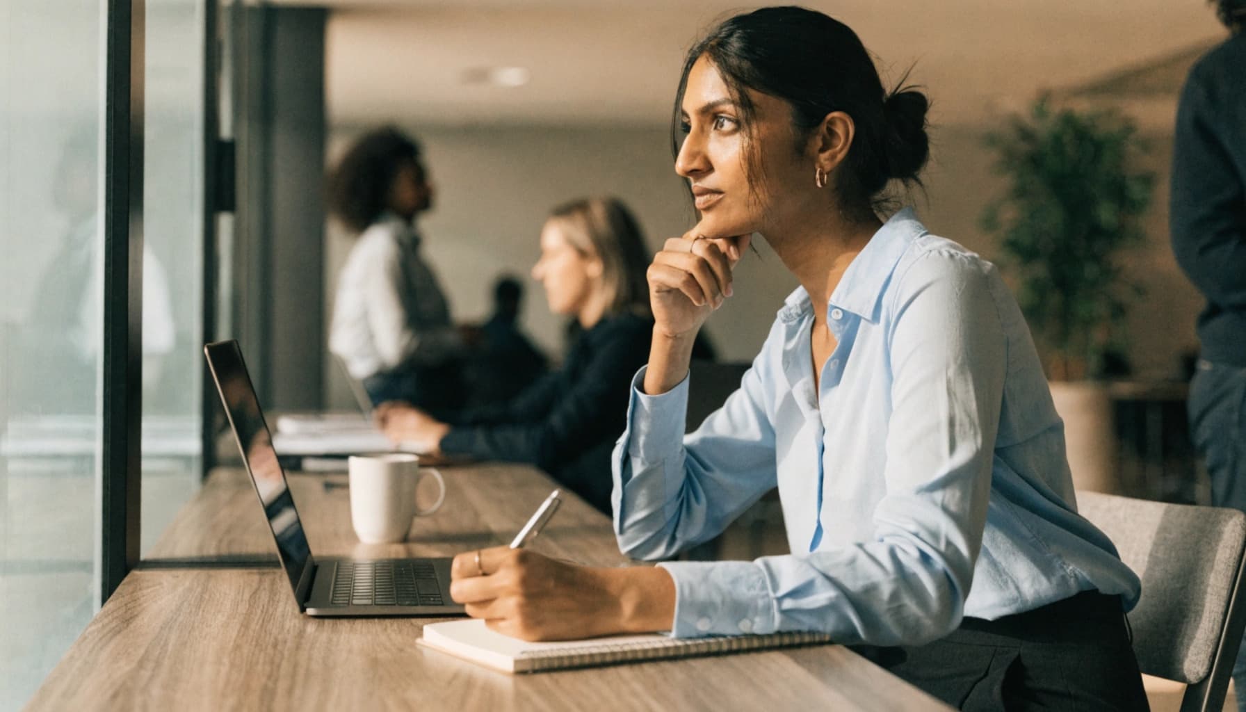 Thoughtful woman in light blue shirt works on a laptop at a bright office desk.