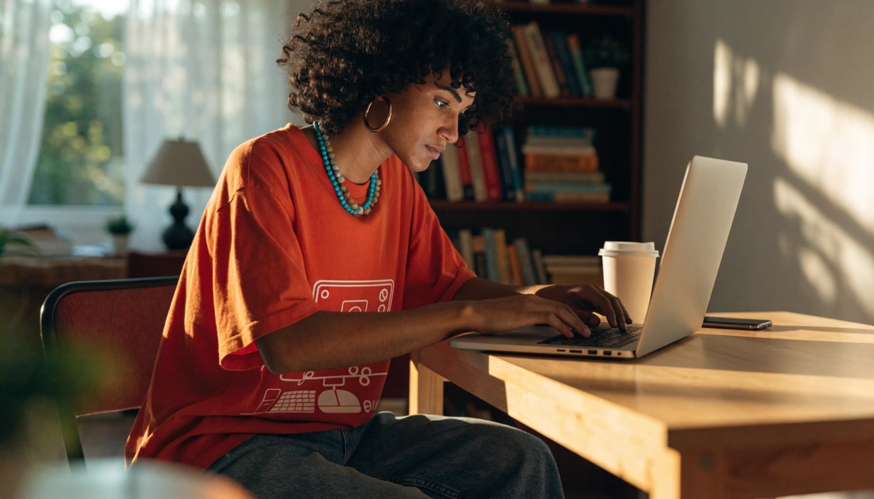 Person with curly hair typing on laptop at wooden desk in sunny room.